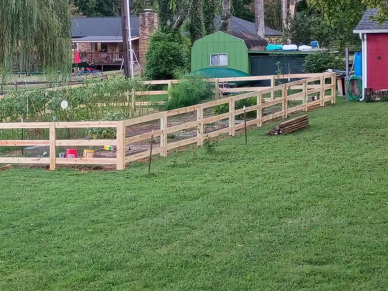 New wood rail fence around a large backyard vegetable garden, with sheds behind it