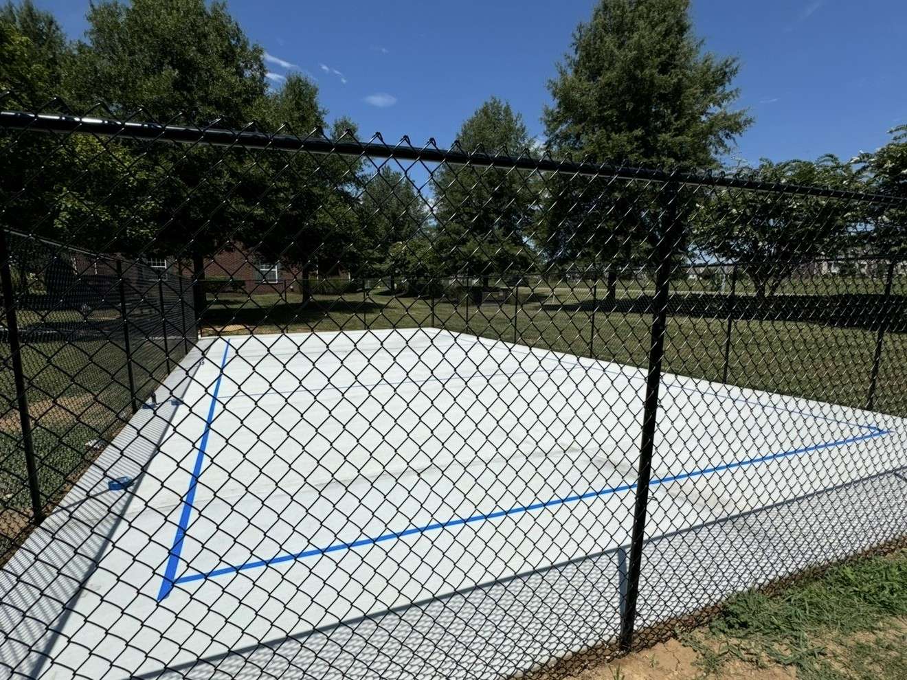 Close view through a new black chain link fence of a concrete court with blue lines, trees behind it