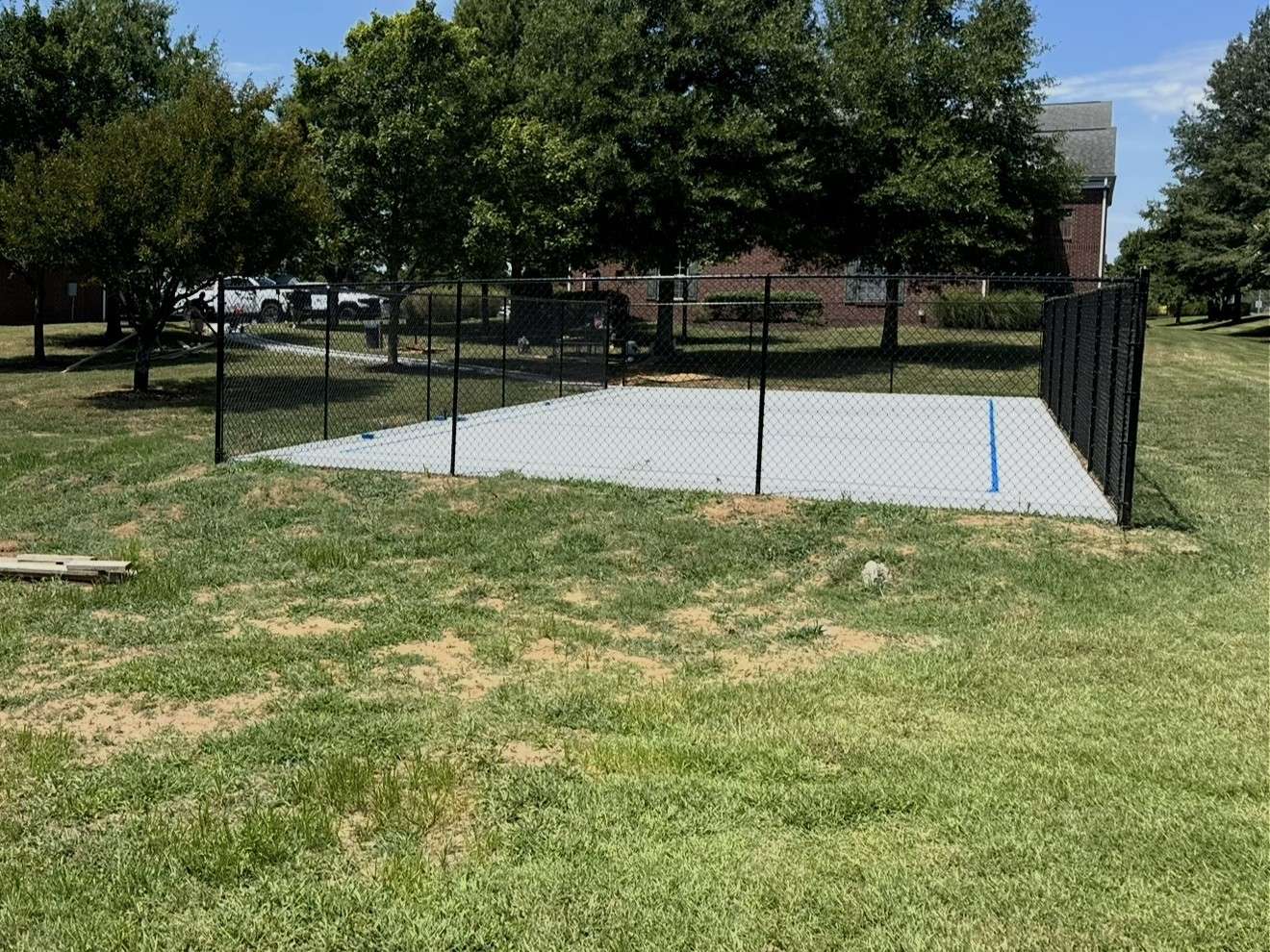 Black chain link fence enclosing a new concrete court on a lawn, with trees and a brick building behind it