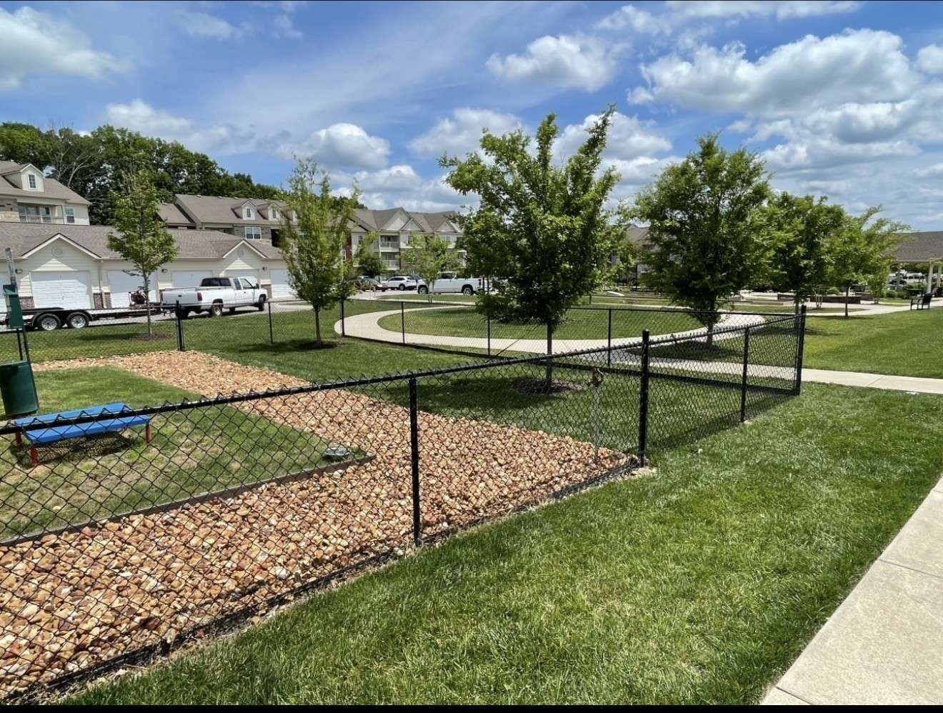 Black chain link fence around a dog park with a gravel strip and a bench, beside a sidewalk at an apartment community