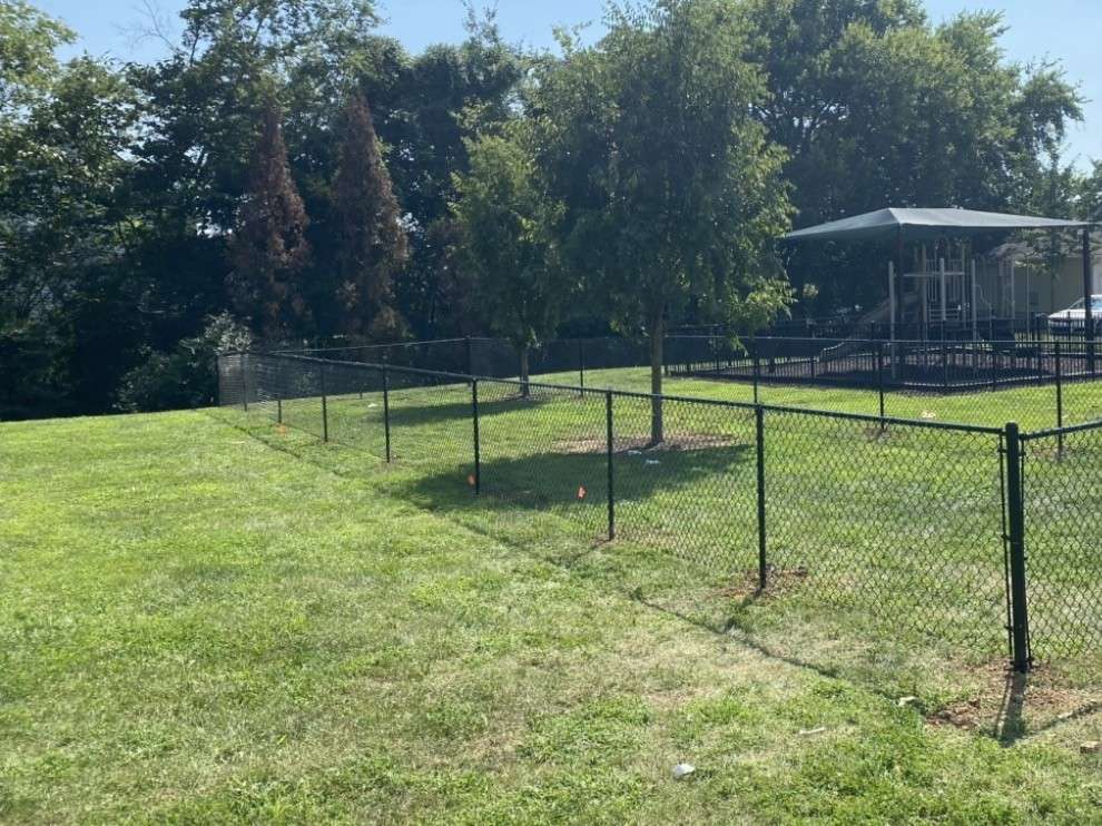 Black chain link fence around a grassy play area with a shaded playground behind it