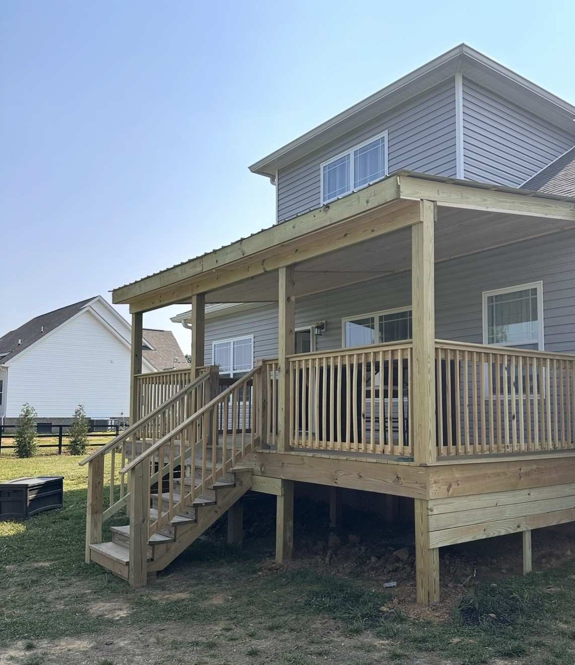Finished raised pressure treated deck with a covered roof, wood railings, and stairs to the yard on the back of a gray two story house