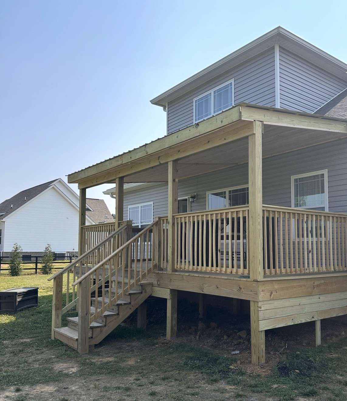 Finished raised pressure treated deck with a covered roof, wood railings, and stairs to the yard on the back of a gray two story house