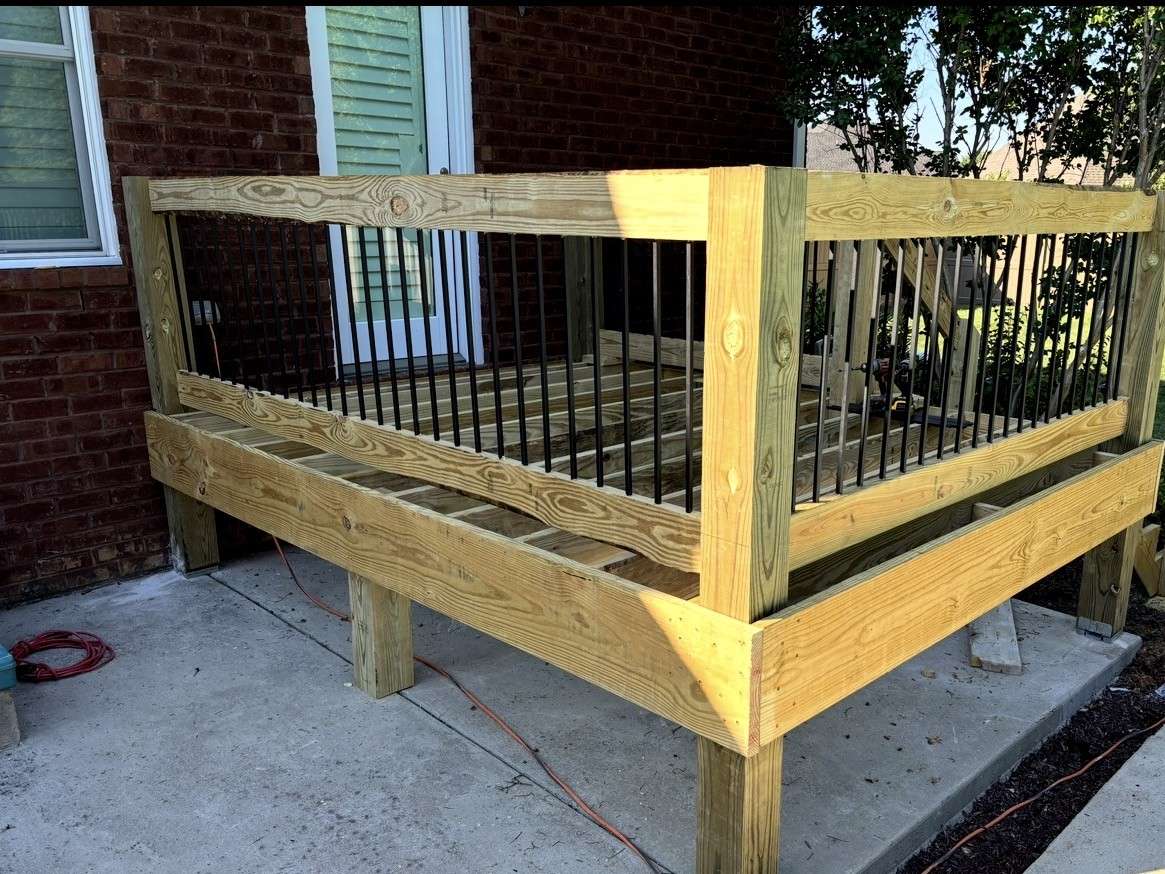 New pressure treated deck frame with wood rails and black metal balusters beside a brick house, before the decking goes down