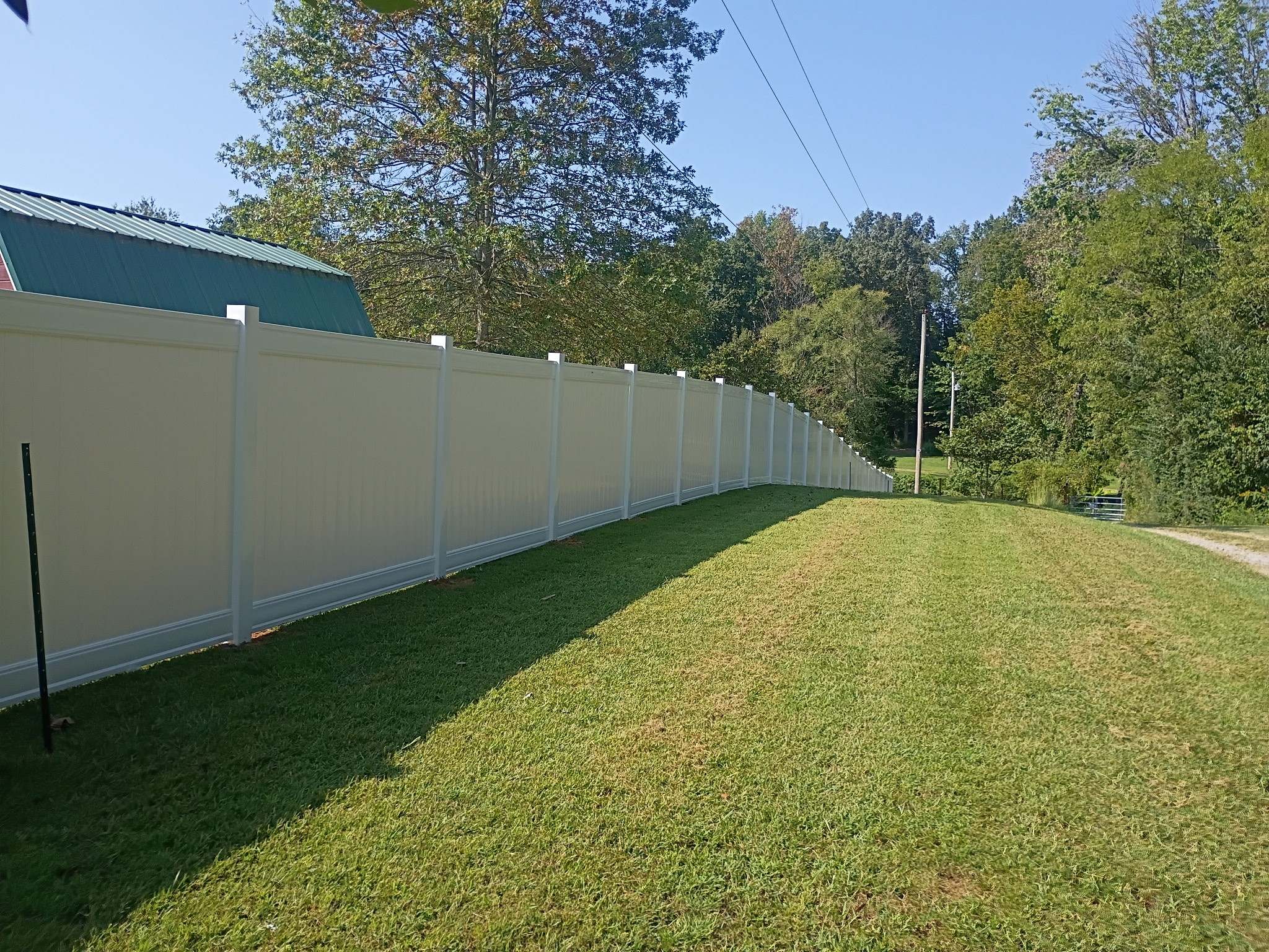 Light colored vinyl privacy fence running along a long sloping fence line beside a barn with a green metal roof